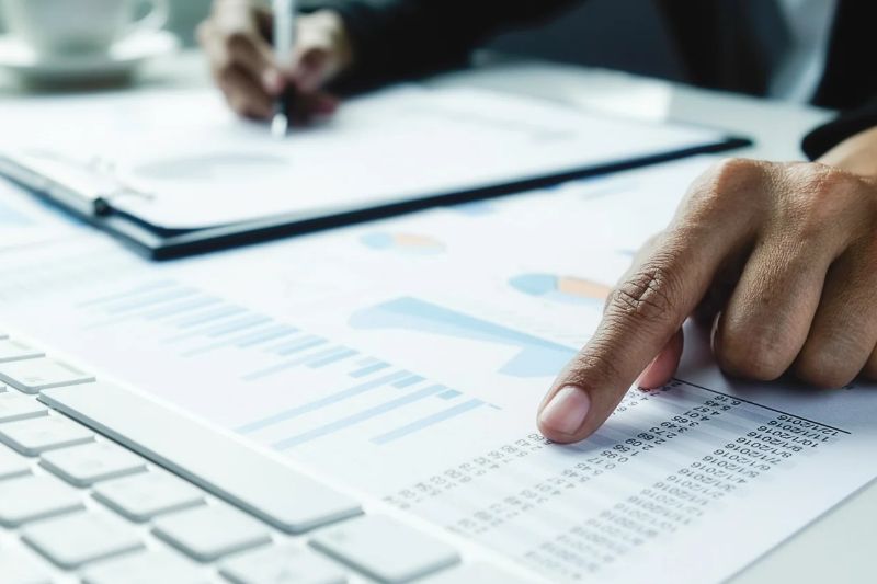 Parte de um teclado de computador, com uma folha embaixo contendo gr&aacute;ficos e n&uacute;meros e uma m&atilde;o apontando pra um dos n&uacute;meros. Do lado desse papel, tem uma prancheta com mais mais gr&aacute;ficos e uma m&atilde;o escrevendo algo