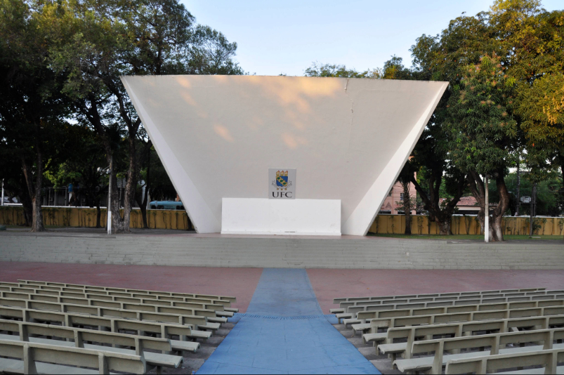 Anfiteatro ao ar livre com palco branco em formato de concha, brasão da UFC ao centro, bancos alinhados e corredor azul. Árvores cercam o espaço, iluminado pela luz suave do fim da tarde