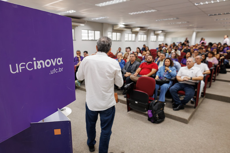 Auditório com plateia cheia durante palestra de um ano da Agência UFC Inova, com um cubo na cor roxa em destaque no lado esquerdo da foto, com a inscrição “ufcinova.ufc.br”