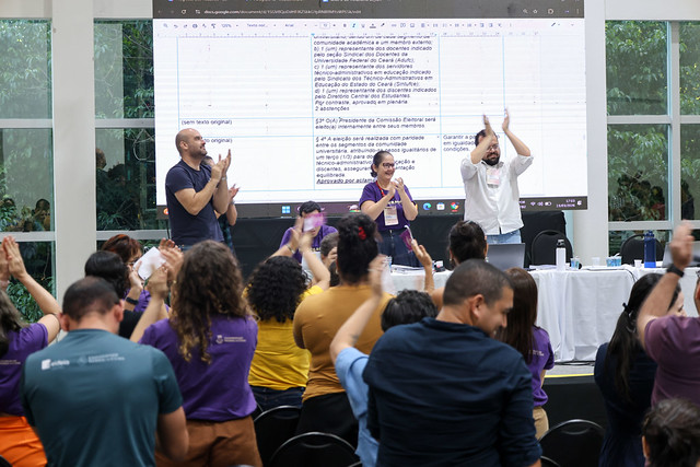 Plateia e palestrantes aplaudem de pé em um auditório. No palco, três pessoas de frente para o público aplaudem diante de um telão que exibe uma tabela com três colunas.