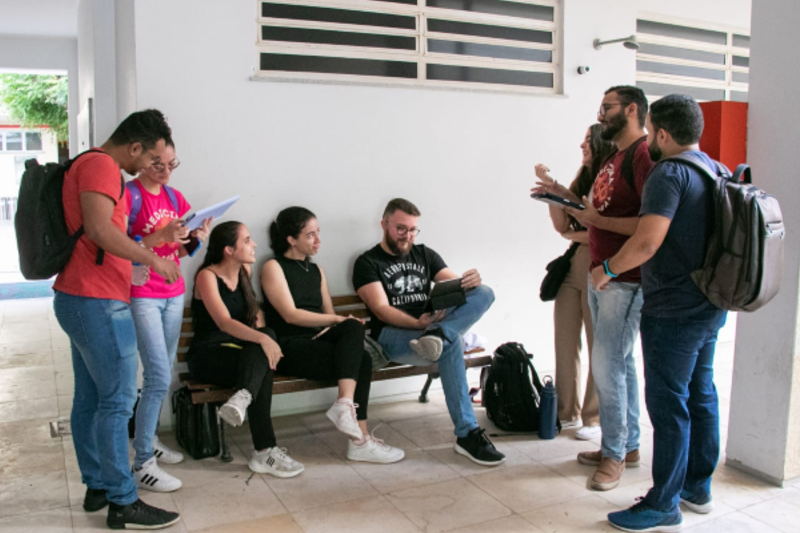 Grupo de estudantes conversa em um corredor da instituição. Alguns estão sentados em um banco e outros em pé, utilizando cadernos, tablet e notebook.