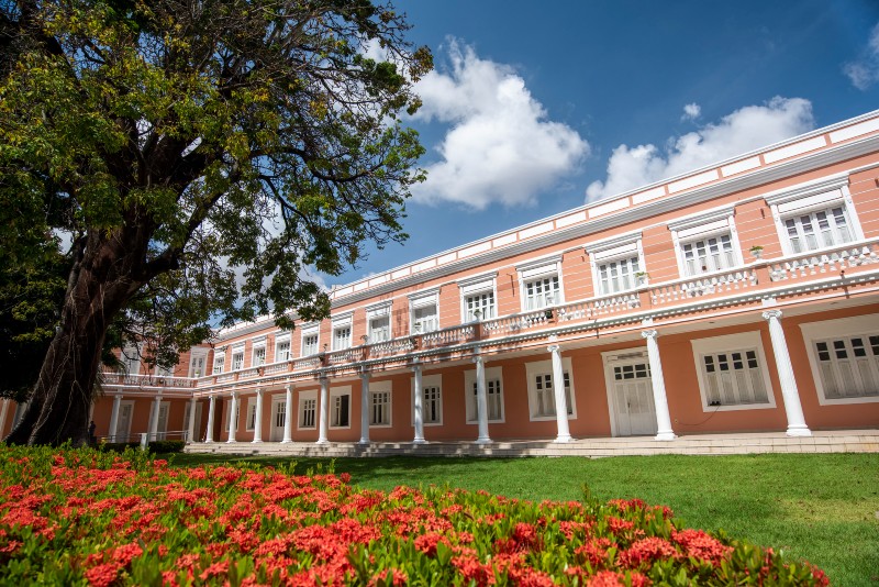 Imagem de edif&iacute;cio hist&oacute;rico em tons rosa, com grandes janelas e colunas. &Agrave; frente, h&aacute; um jardim florido, gramado verde e uma grande &aacute;rvore, sob c&eacute;u azul com nuvens