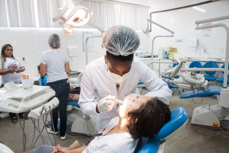 Foto mostra uma mulher branca, sentada em cadeira odontológica, recebendo atendimento. A dentista é mulher, negra, e está de jaleco, luvas, máscara e touca. Ao fundo, aparecem outras pessoas e equipamento odontológico (Foto: Ribamar Neto/UFC)