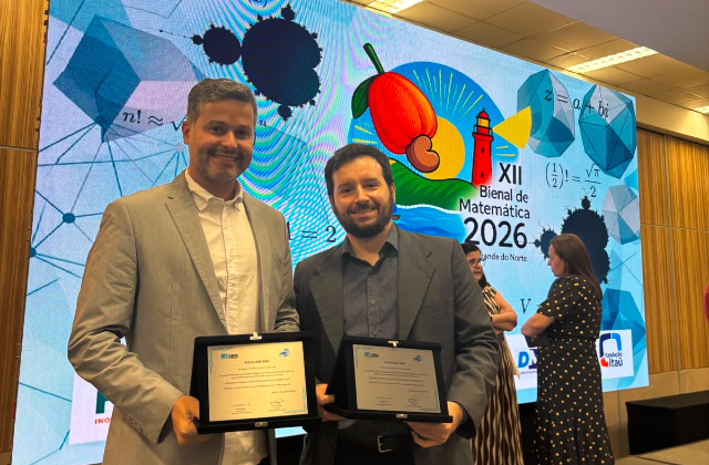 Foto mostra dois professores segurando certificados de premia&ccedil;&atilde;o, em frente a um banner onde se pode ler: XII Bienal da Matem&aacute;tica (foto: Acervo pessoal)