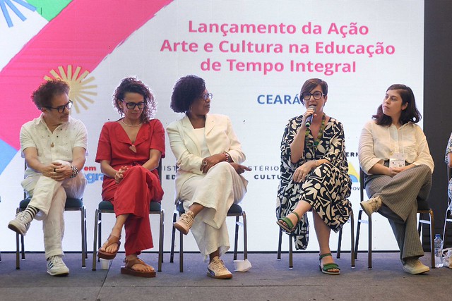 ft_260401_culturatempointegral_gr Cinco pessoas sentadas em cadeiras sobre um palco durante uma mesa de debate. Ao fundo, uma tela exibe informações sobre um evento de arte e cultura na educação.
