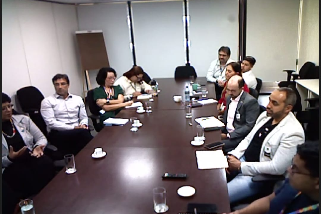 Cerca de dez pessoas participam de uma reunião em uma sala institucional, sentadas ao redor de uma mesa com documentos, blocos de anotação e bebidas. Uma participante fala enquanto os demais acompanham a discussão