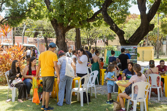 Pessoas participam de atividade coletiva ao ar livre, reunidas em mesas distribu&iacute;das sobre &aacute;rea gramada. Adultos e crian&ccedil;as conversam, escrevem e interagem em grupo sob &aacute;rvores.