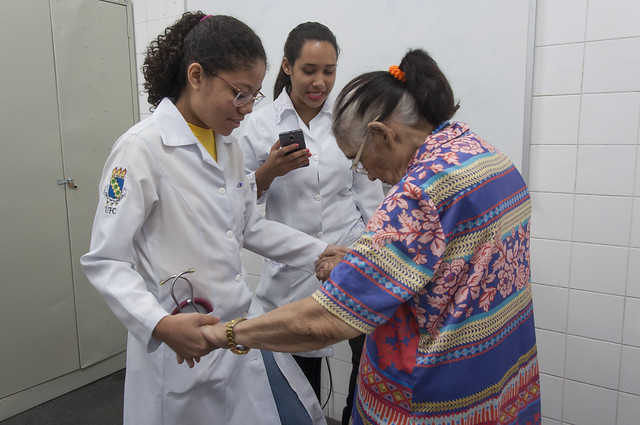 Imagem: uma jovem de jaleco branco segura uma mulher idosa pelas mãos. As duas estão de pé. Uma outra jovem, também de jaleco branco, aponta um celular para as duas