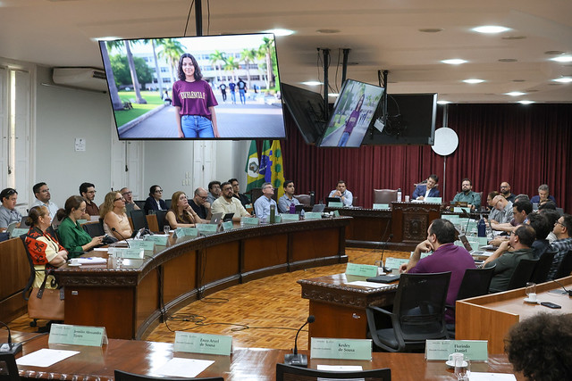 Pessoas reunidas em torno de uma grande mesa curva de madeira em um plenário. Telas suspensas no teto exibem a foto de uma jovem em um campus. Ao fundo, veem-se bandeiras oficiais e uma cortina vermelha.