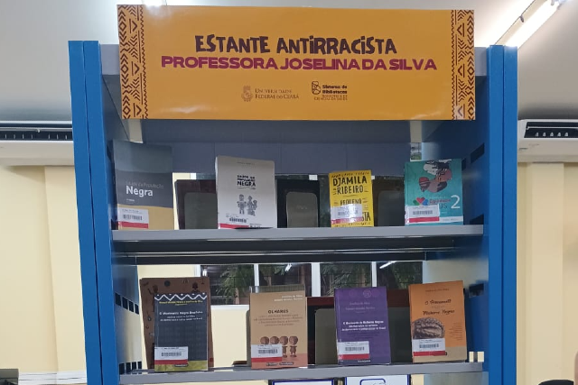 Estante azul com livros em exposi&ccedil;&atilde;o sob uma placa amarela onde se l&ecirc;: &ldquo;Estante Antirracista Professora Joselina da Silva&rdquo;.