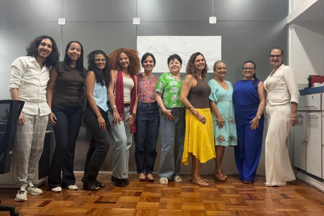 Grupo de dez pessoas (nove mulheres e um homem) posa sorrindo em sala com piso de madeira e parede cinza.