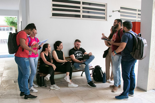 Oito estudantes de roupas casuais conversam e estudam em um corredor de prédio universitário com paredes brancas e piso claro.