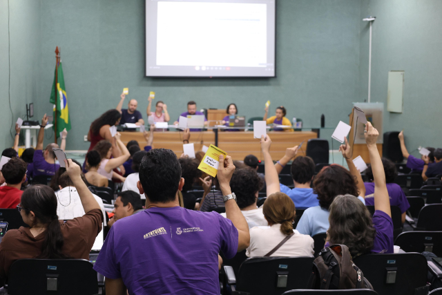 Participantes sentados em auditório, muitos de camiseta roxa, erguem cartões roxos em votação. No centro, um homem de barba observa.