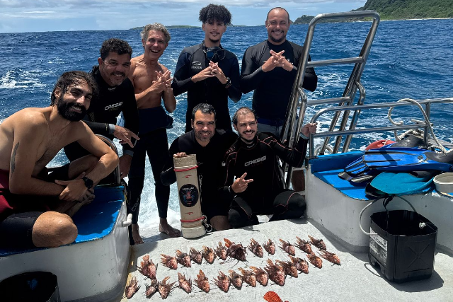 ft_260515_equipepeixeleao_gr Sete pessoas da equipe de mergulho posam sorrindo em um barco com vários peixes-leão alinhados no piso. Ao fundo, o mar.