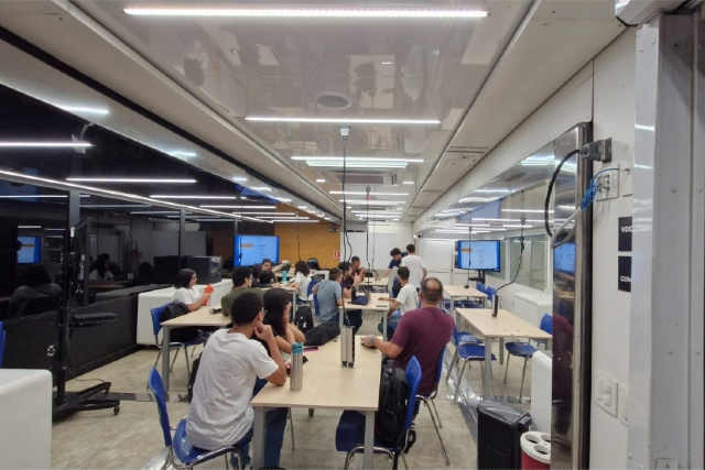 ft_260519_caminhaodentro_gr Imagem: imagem de dentro de uma sala de aula com mesas, cadeiras, estudantes de costas olhando pra uma lousa e sentados ao redor de mesas. Local com janelas de vidros.
