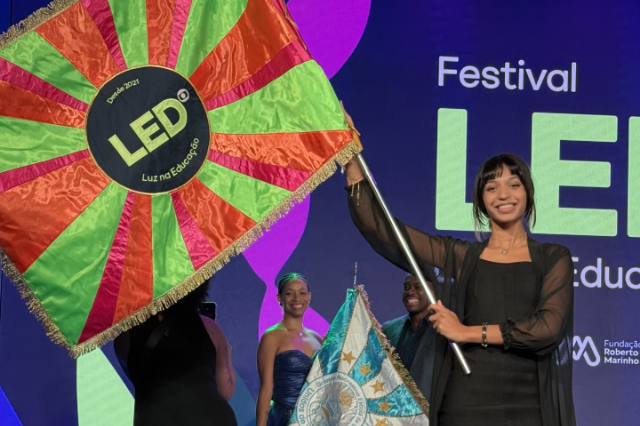 Jenifer, sorridente, segura bandeira listrada em verde e rosa com a marca "LED". Ao fundo, painel do Festival LED.