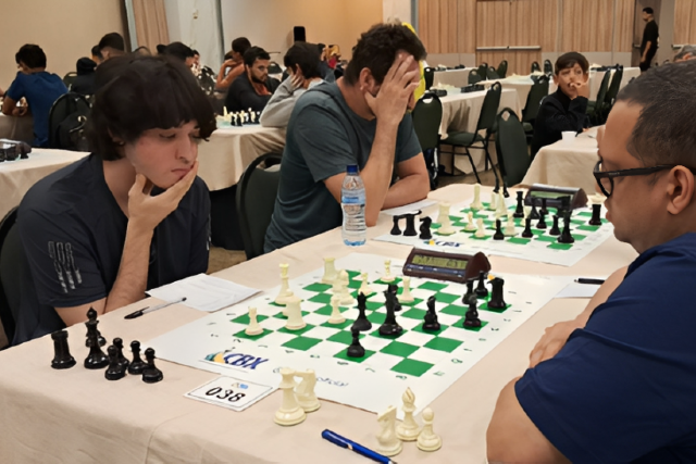 Três enxadristas concentrados jogam uma partida de xadrez em um salão amplo onde ocorre um torneio.