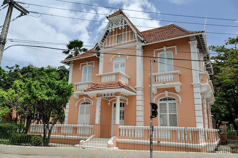 Descrição: Fachada da Casa de Cultura Alemã da UFC. Casarão histórico de dois andares, na cor salmão com detalhes brancos e telhado avermelhado. Entrada com colunas, cercada por grades e árvores.