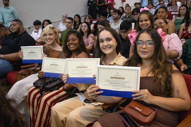 Auditório durante uma cerimônia acadêmica. Em primeiro plano, quatro jovens sentadas lado a lado sorriem para a câmera enquanto seguram certificados. Elas usam roupas em tons claros e terrosos."