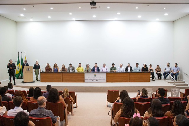 auditório durante cerimônia acadêmica. No palco, pessoas estão sentadas atrás de uma longa mesa, compondo a mesa de honra. À esquerda, uma pessoa fala ao microfone em um púlpito. Na plateia, participantes acompanham evento