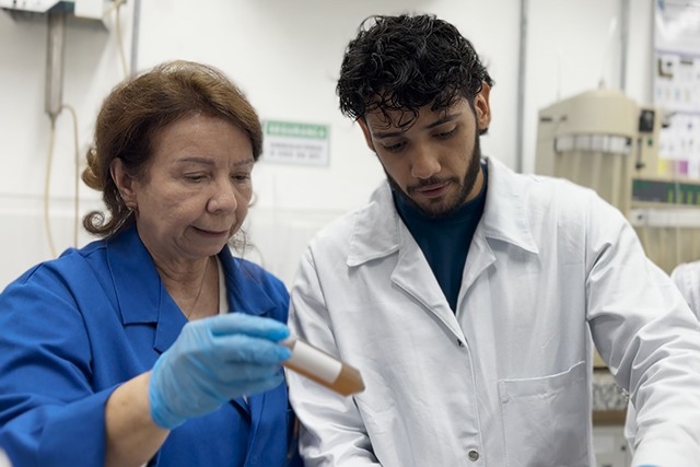 Mulher de jaleco azul e luva segura tubo de ensaio com líquido âmbar, enquanto um jovem de jaleco branco observa atentamente ao seu lado em um laboratório. Ao fundo, equipamentos científicos desfocados.