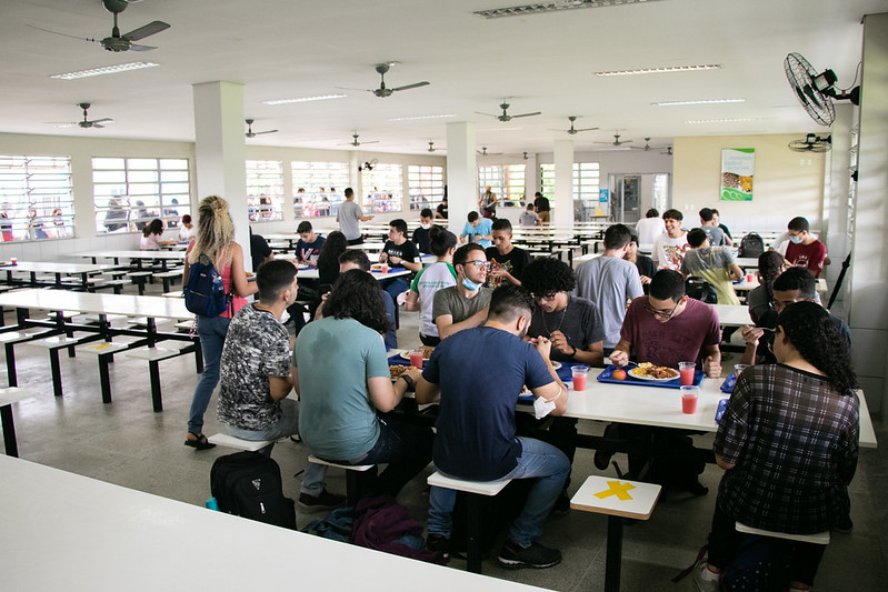 Foto interna do Restaurante Universitário do Campus do Pici da UFC, com mesa à direita ocupada por nove estudantes, uma aluna de blusa rosa e mochila preta caminhando no corredor e carregando uma bandeja, e mais três mesas ao fundo do refeitório, à direit
