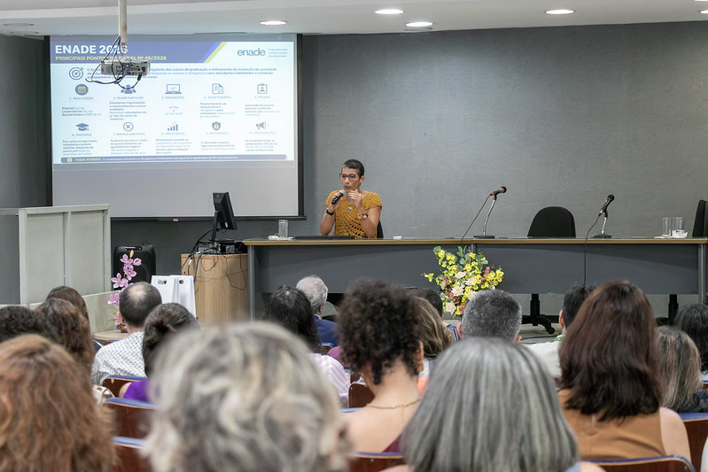 Foto mostra pessoas sentadas nas cadeiras do auditório e um palco com uma mesa onde está sentada Ana Júlia, coordenadora-geral de Avaliação das Licenciaturas do Inep. Ao lado direito dela, vê-se uma grande tela com informações sobre o Enade