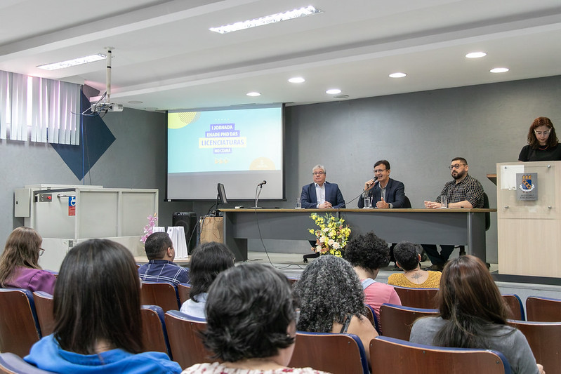 foto de pessoas sentadas nas cadeiras do auditório e, à frente delas, um palco com uma mesa onde estão sentados, da esquerda para a direita, Davi Romero, Custódio Almeida e Cícero Miranda