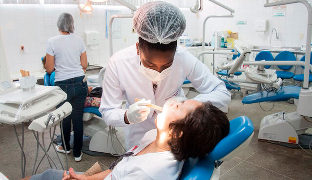 Profissional de odontologia, usando máscara, luvas e touca, realiza exame bucal em uma paciente reclinada em uma cadeira odontológica. Ao fundo, há outros consultórios e pessoas em atendimento em uma clínica odontológica