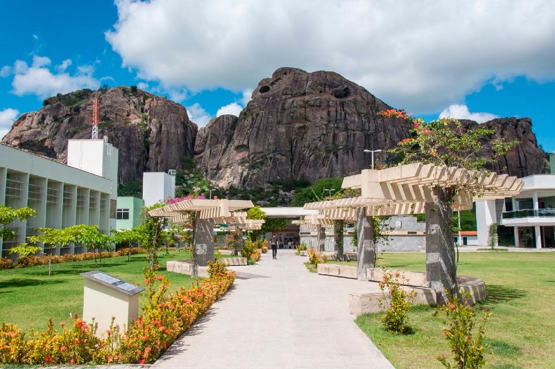 Trata-se de uma foto do Campus da UFC em Quixadá. Em primeiro plano, um caminho arborizado com pergolados e jardins leva aos prédios da universidade. Ao fundo, destacam-se os grandes monólitos de Quixadá sob céu azul com nuvens brancas.