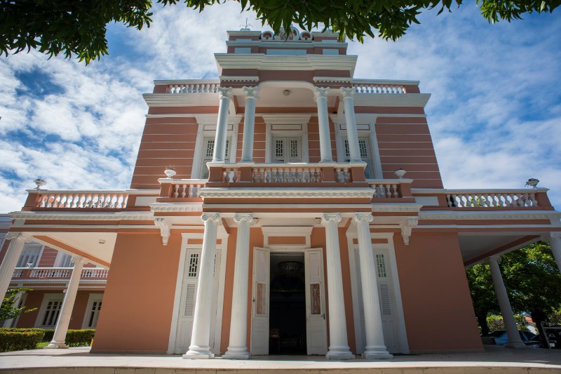 Trata-se de uma foto da entrada do Auditório da Reitoria da Universidade Federal do Ceará (UFC). A imagem destaca a fachada histórica do edifício, com colunas brancas, portas abertas e elementos arquitetônicos clássicos emoldurados pelo céu e pela ve