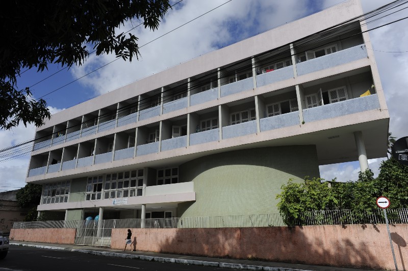 Prédio de vários andares, com fachada branca e azul, amplas varandas e muitas janelas. Em frente, há muro, grade, árvores, fios elétricos e uma pessoa caminhando pela calçada