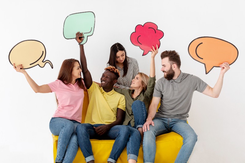 Trata-se de uma foto de cinco pessoas sentadas em um sof&aacute; amarelo, sorrindo e segurando bal&otilde;es coloridos em formato de di&aacute;logo. Ao fundo, uma parede branca, sugerindo comunica&ccedil;&atilde;o, troca de ideias e trabalho em equipe.