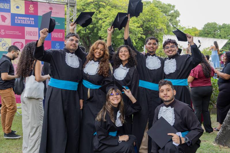Grupo de sete estudantes, homens e mulheres, vestindo beca de formatura e levantando o capelo, nos jardins da Reitoria da UFC