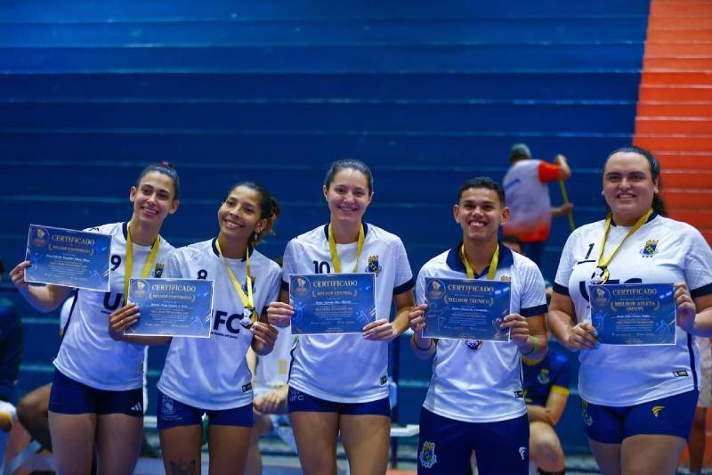 Quatro atletas mulheres e um homem vesidos de uniforme de v&ocirc;lei, lado a lado, segurando medalhas e um certificado do campeonato