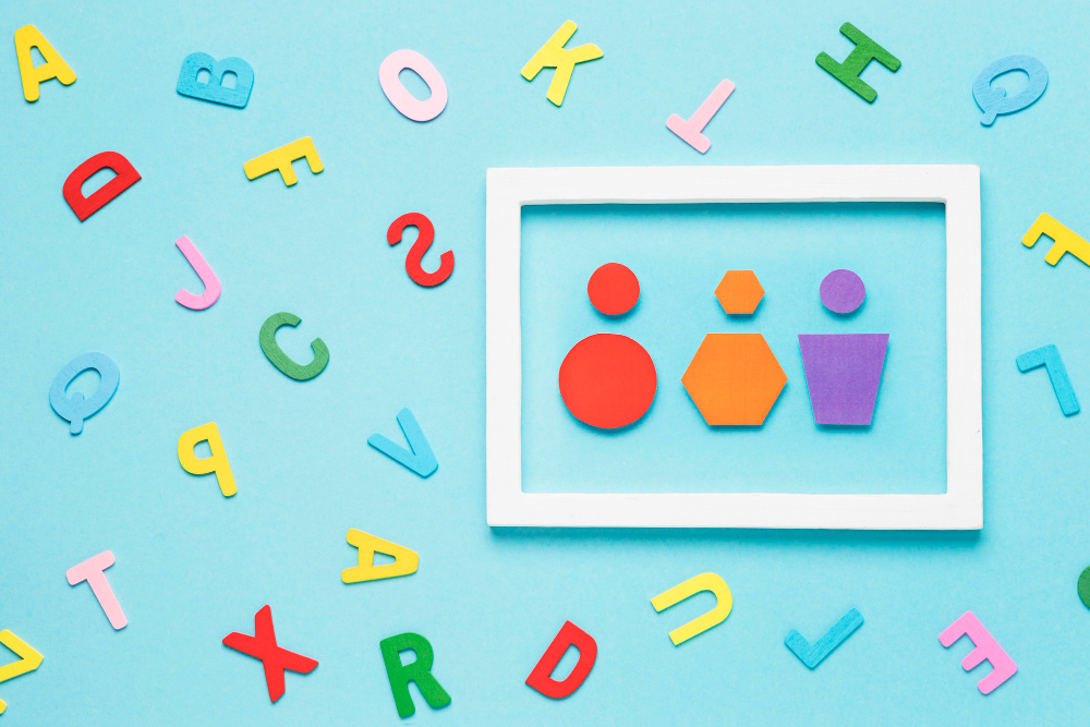 Imagem com fundo azul-claro, letras e formas geom&eacute;tricas coloridas espalhadas ao redor de uma moldura branca. Dentro dela, c&iacute;rculos, hex&aacute;gonos e um trap&eacute;zio comp&otilde;em uma composi&ccedil;&atilde;o l&uacute;dica e educativa.