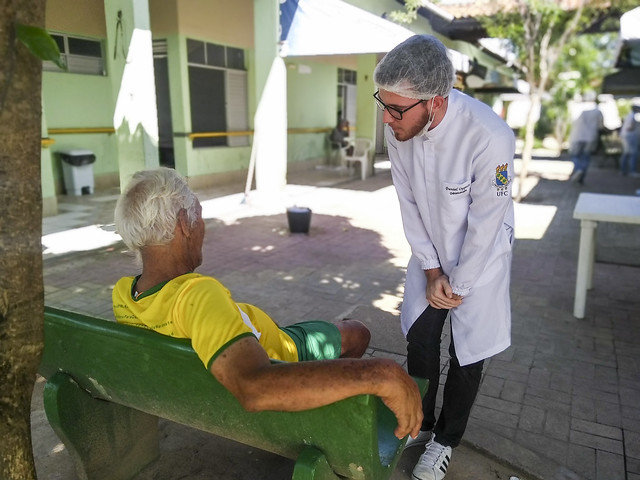 Uma pessoa idosa de camisa amarela sentada em um banco de cor verde; diante dela, um jovem com jaleco branco e marca da UFC na manga, conversando com o idoso