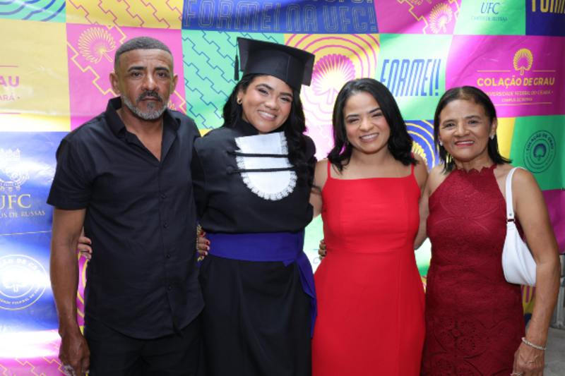 Foto de quatro pessoas em uma cerim&ocirc;nia de cola&ccedil;&atilde;o de grau. Ao centro, uma mulher usa beca e capelo, acompanhada por um homem, uma mulher de vestido vermelho e outra de vestido vinho.