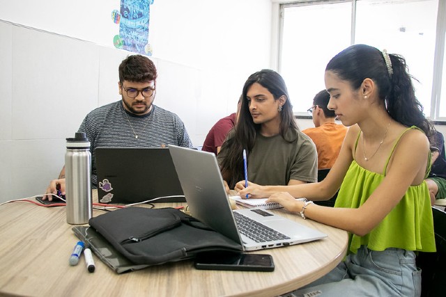 Dois alunos e uma aluna sentados em uma mesa redonda escrevendo em papéis, com um notebook, garrafa e canetas sobre a mesa