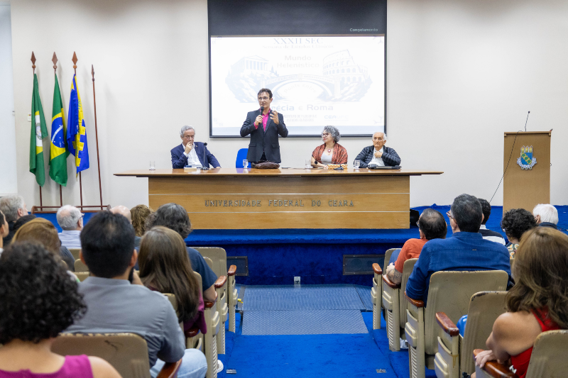 Foto da mesa do evento com o reitor Cust&oacute;dio Almeida em p&eacute;, os convidados sentados e o p&uacute;blico no primeiro plano