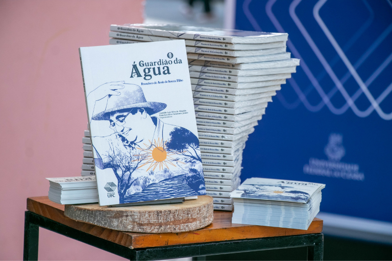 Foto de uma mesa com exemplares empilhados do livro "O Guardi&atilde;o da &Aacute;gua" no estande de vendas da Editora UFC