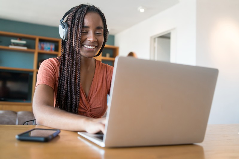 Uma mulher negra, sorridente, usa fones de ouvido enquanto trabalha em um notebook sobre uma mesa de madeira. Ao fundo, h&aacute; uma estante com livros e objetos decorativos em um ambiente residencial iluminado