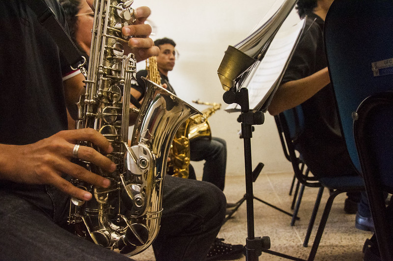 A imagem mostra alguns músicos vestido de camisa preta e tocando instrumentos de sopro