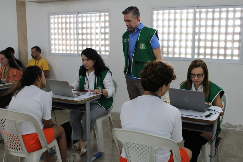 Atendimento em uma sala, com quatro pessoas sentadas e tr&ecirc;s profissionais usando coletes verdes, trabalhando em computadores e orientando os participantes.
