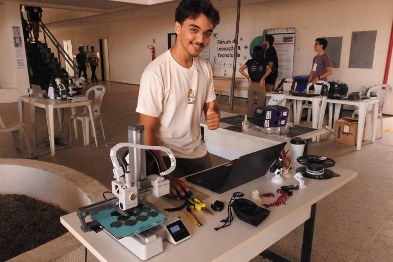 Estudante, vestido de camiseta branca, de frente para uma mesa sobre a qual h&aacute; um computador e equipamentos de texnologia