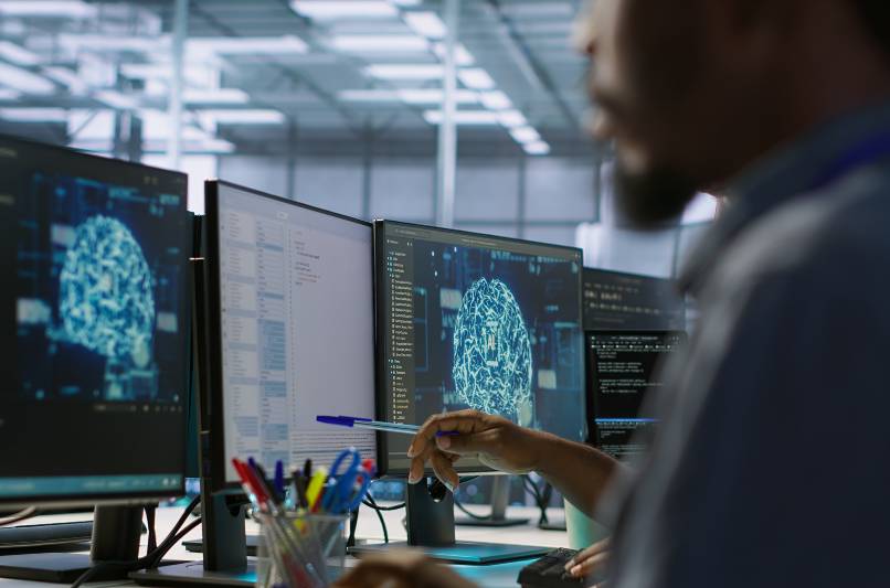 Homem negro visto de perfil, diante de telas de computador com imagens referentes &agrave; intelig&ecirc;ncia artificial