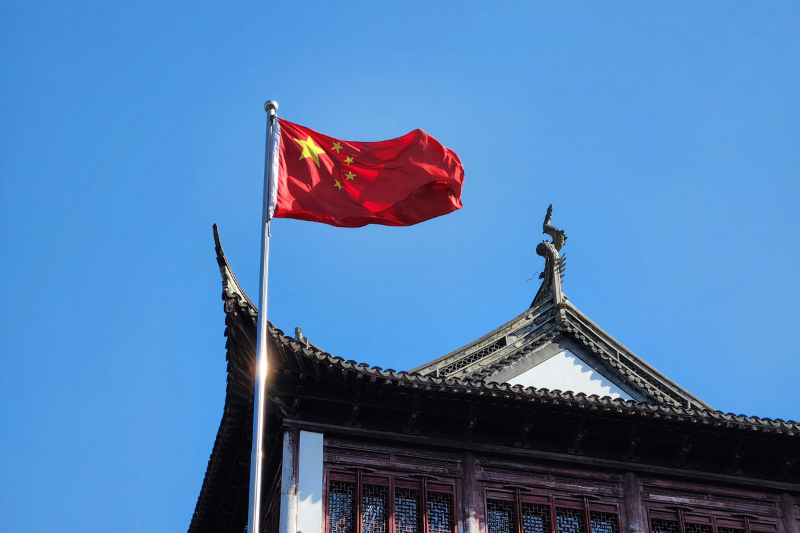 Bandeira da China com fundo vermelho e estrelas amarelas tremulando perto de um pr&eacute;dio hist&oacute;rico no estilo arquitet&ocirc;nico asi&aacute;tico