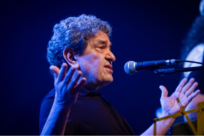 Fausto Nilo vestido de preto, est&aacute; no palco diante de um microfone. Ele gesticula com as duas m&atilde;os enquanto fala ou canta.