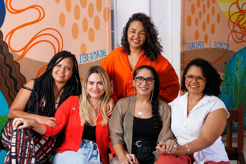 Cinco mulheres posam sorrindo diante de um mural colorido, com desenhos abstratos e as palavras &ldquo;liberdade&rdquo;, &ldquo;igualdade&rdquo; e &ldquo;respeito&rdquo;