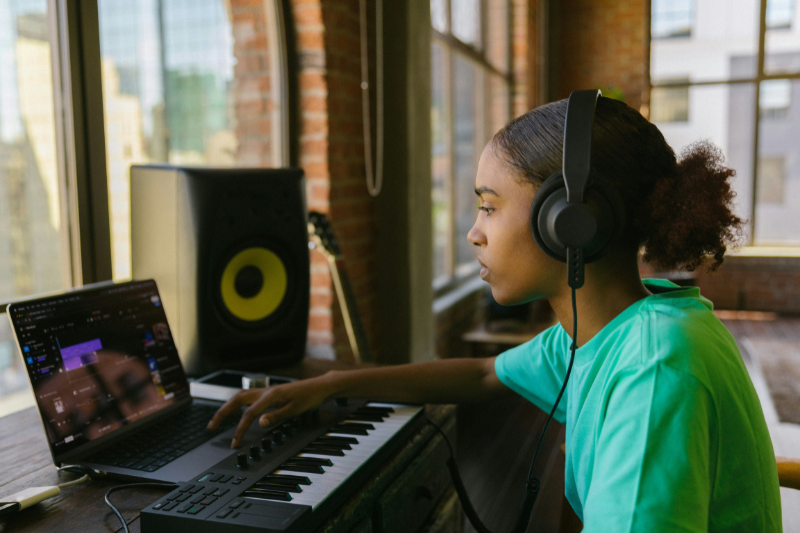 Moça negra que está utilizando fones de ouvido, um computador e um teclado para ouvir as produções de um estúdio de música